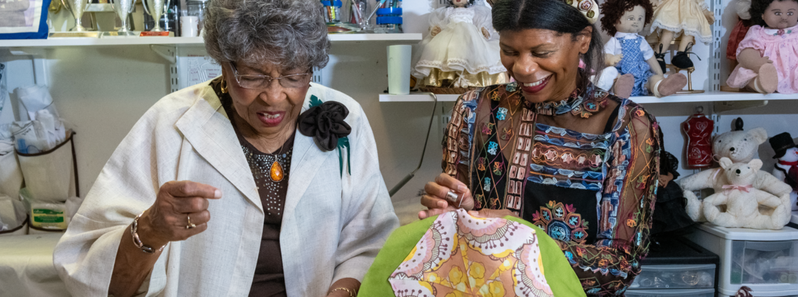 Two people are sewing in front of a wall with dolls on the shelves.